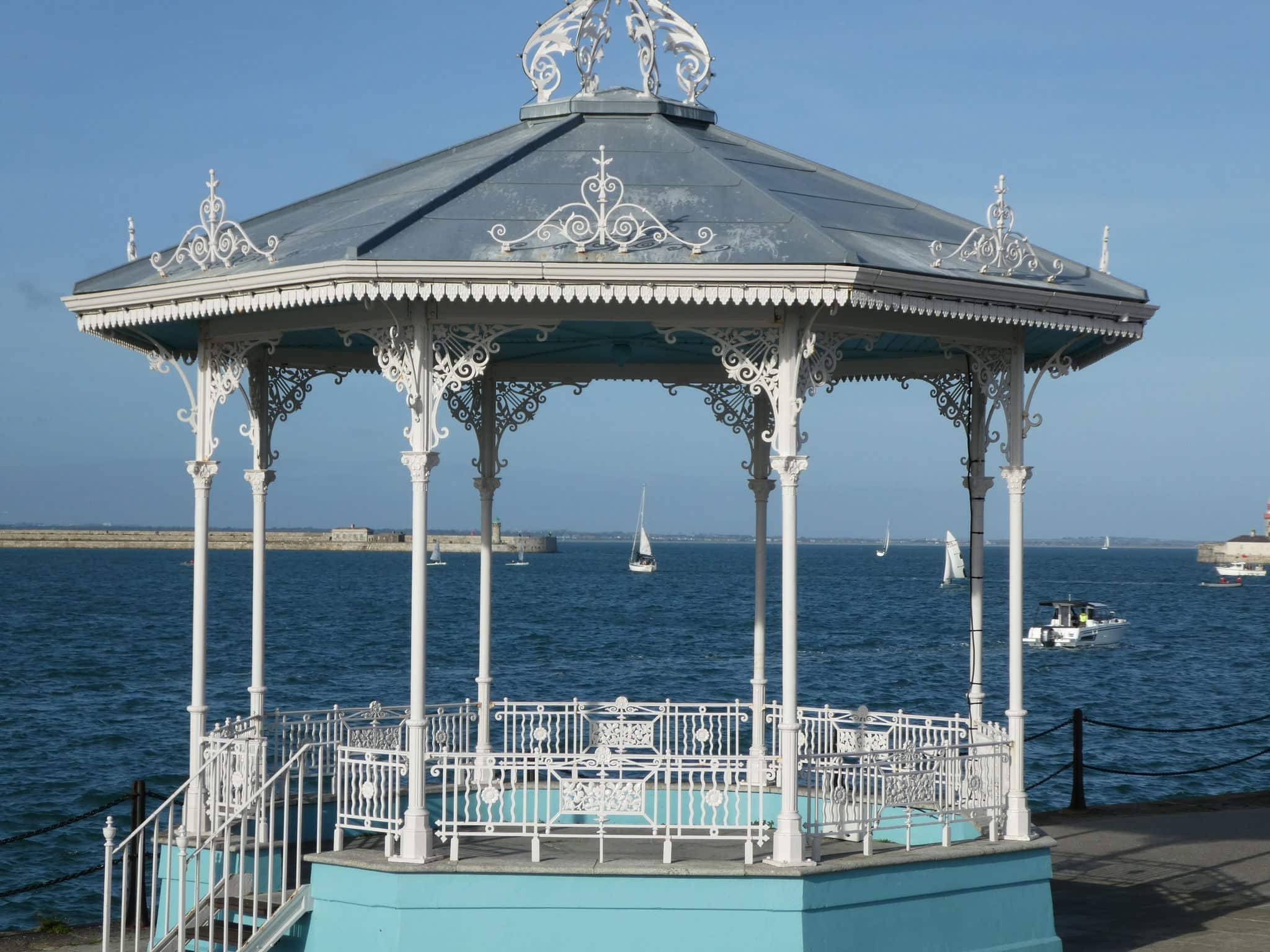 A pleasure yacht can be seen framed by the old white iron work of the band stand at Dun Laoghaire against a bright blue sky and deep blue water.