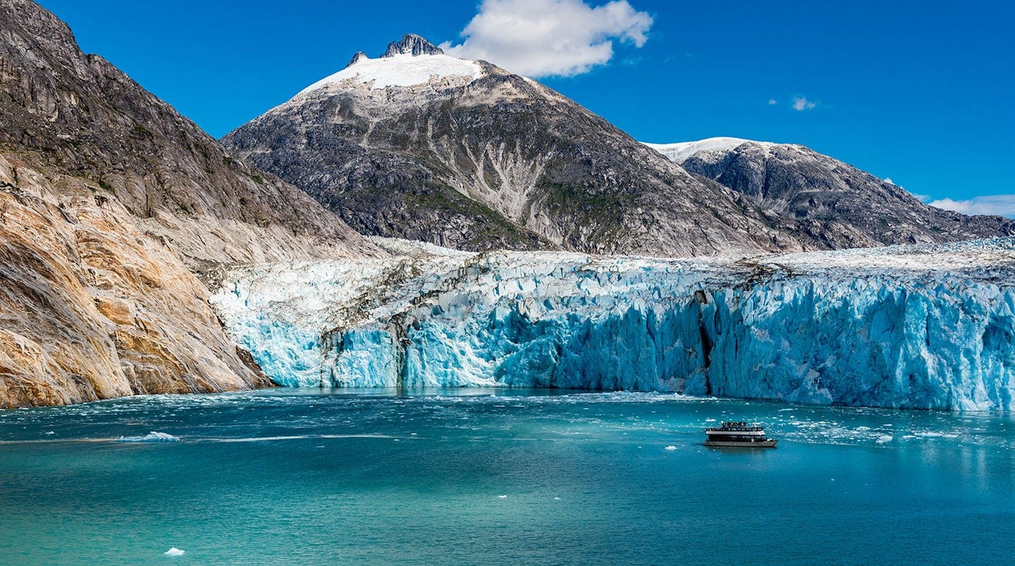 Wide angle view of glacier face with tourist boat and snow capped mountains and dappled sunshine on the icy waters