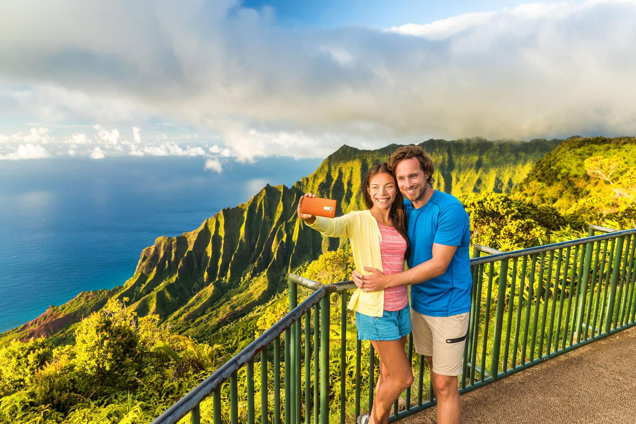 Hawaii travel Napali selfie couple at Napali nature Kauai Hawaii. Young tourists taking selfie phone photo at Na Pali lookout in Kauai, Hawaii. Travel people at famous Kalalau hawaiian attraction.