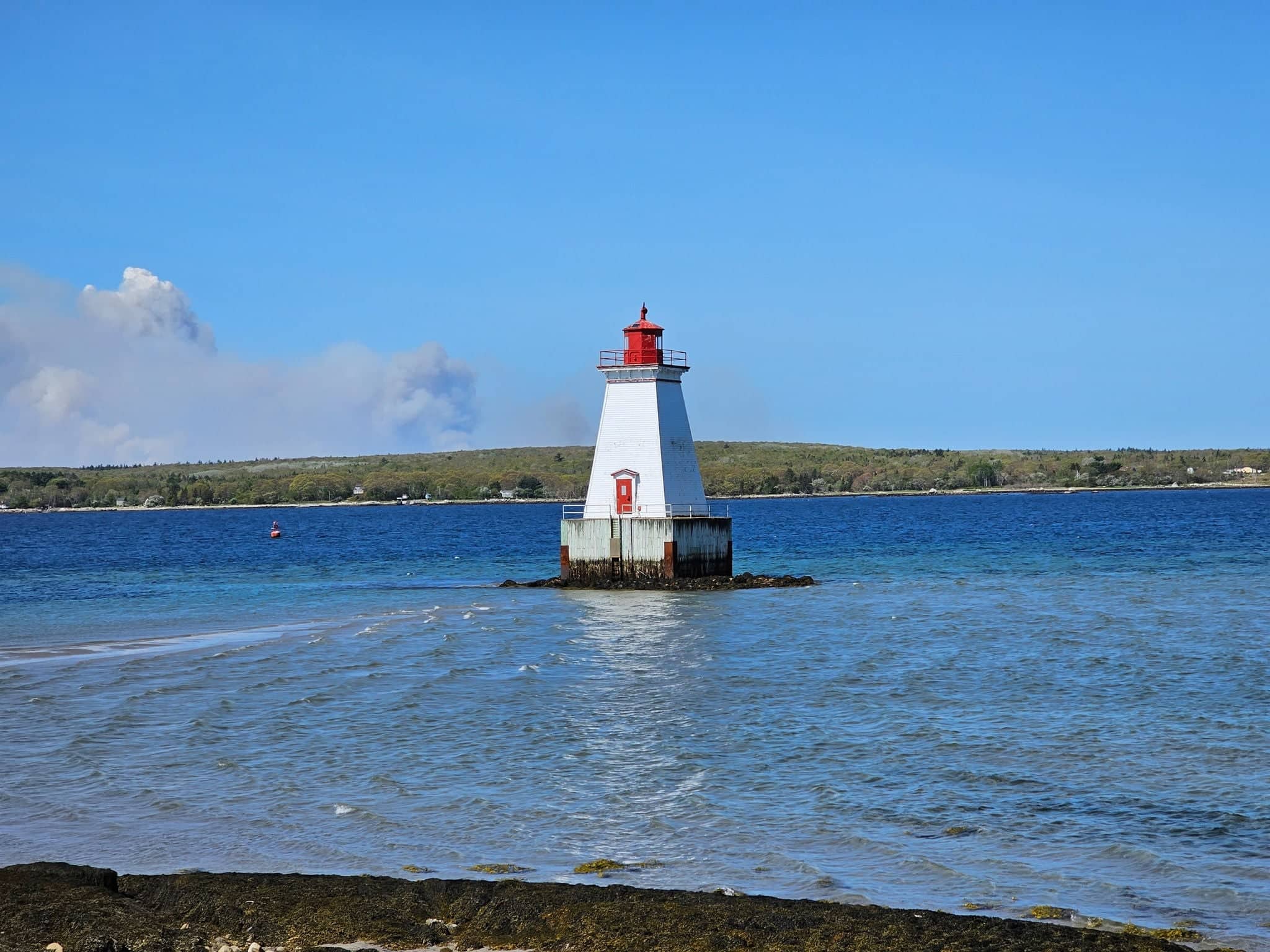 A lighthouse sitting in the middle of the water with a smoke cloud from a wildfire off to the side.