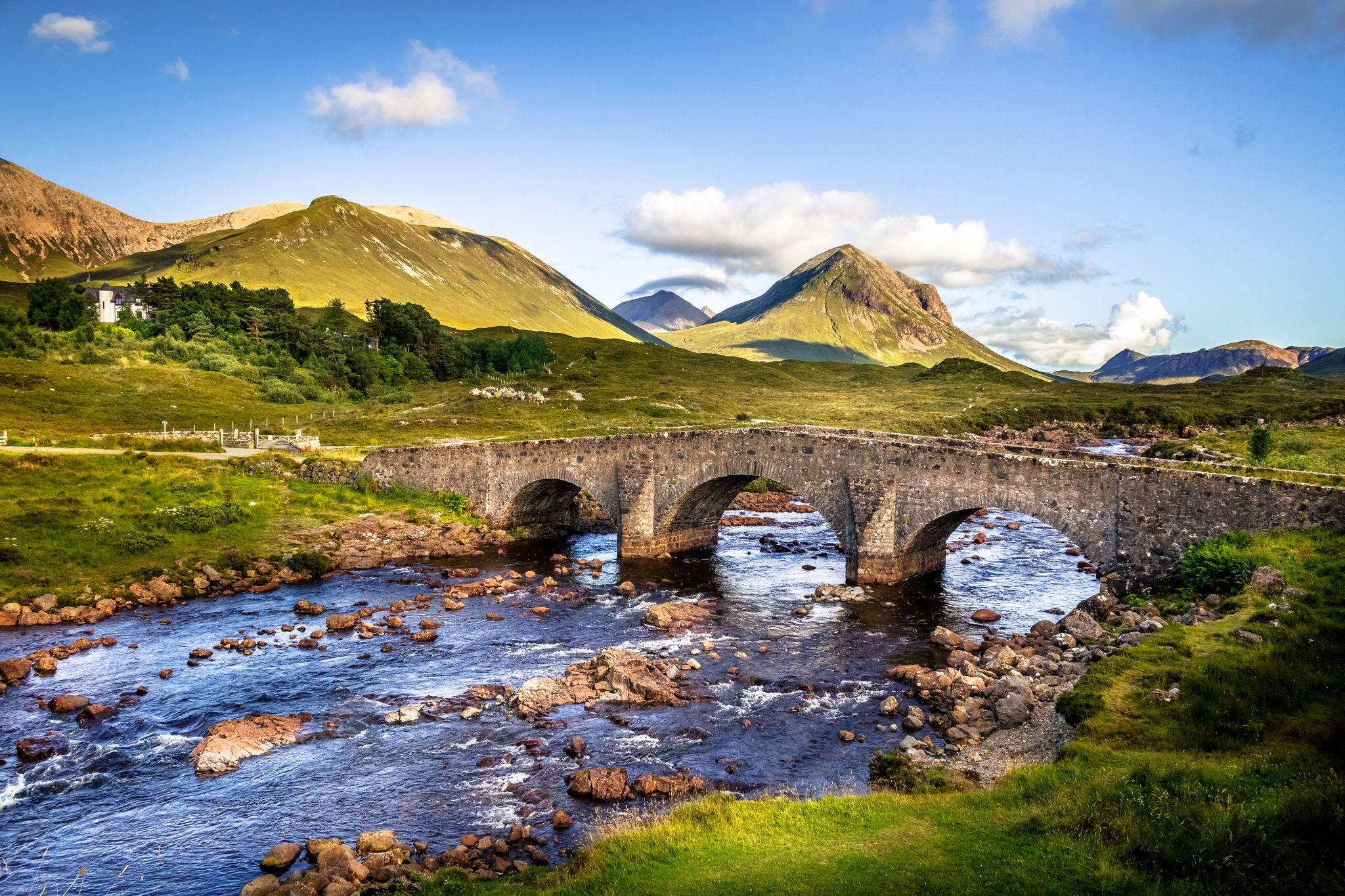Old vintage brick bridge crossing river in Sligachan, Isle of Skye, Scotland with scottish landscape, vegetation, hills and mountains with fresh, blue water and sunny sky