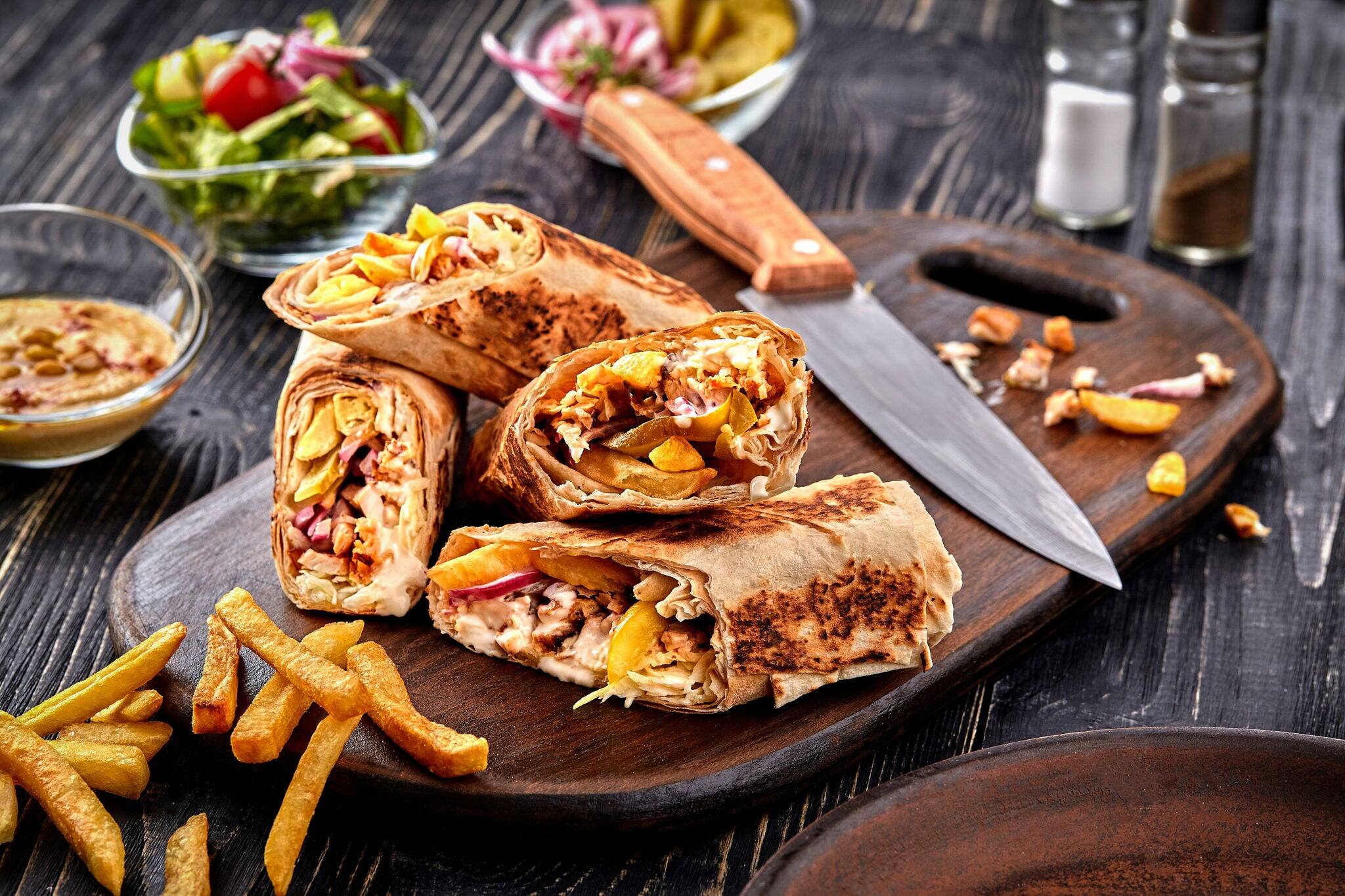 Shawarma chicken roll in a pita with fresh vegetables, cream sauce and french fries on wooden background. Selective focus