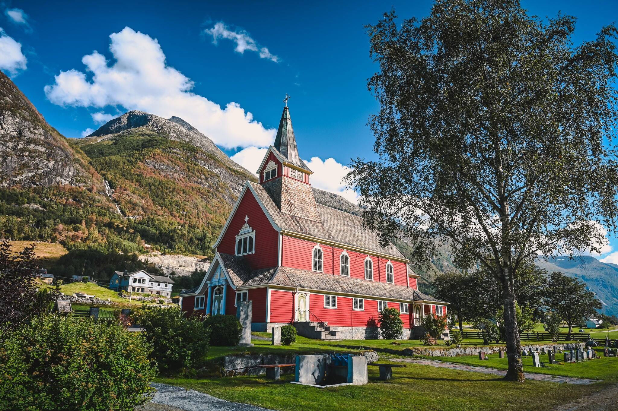 View of the red Olden Church in the village of Olden, Norway on a sunny summer day. Forested mountains are in the background.