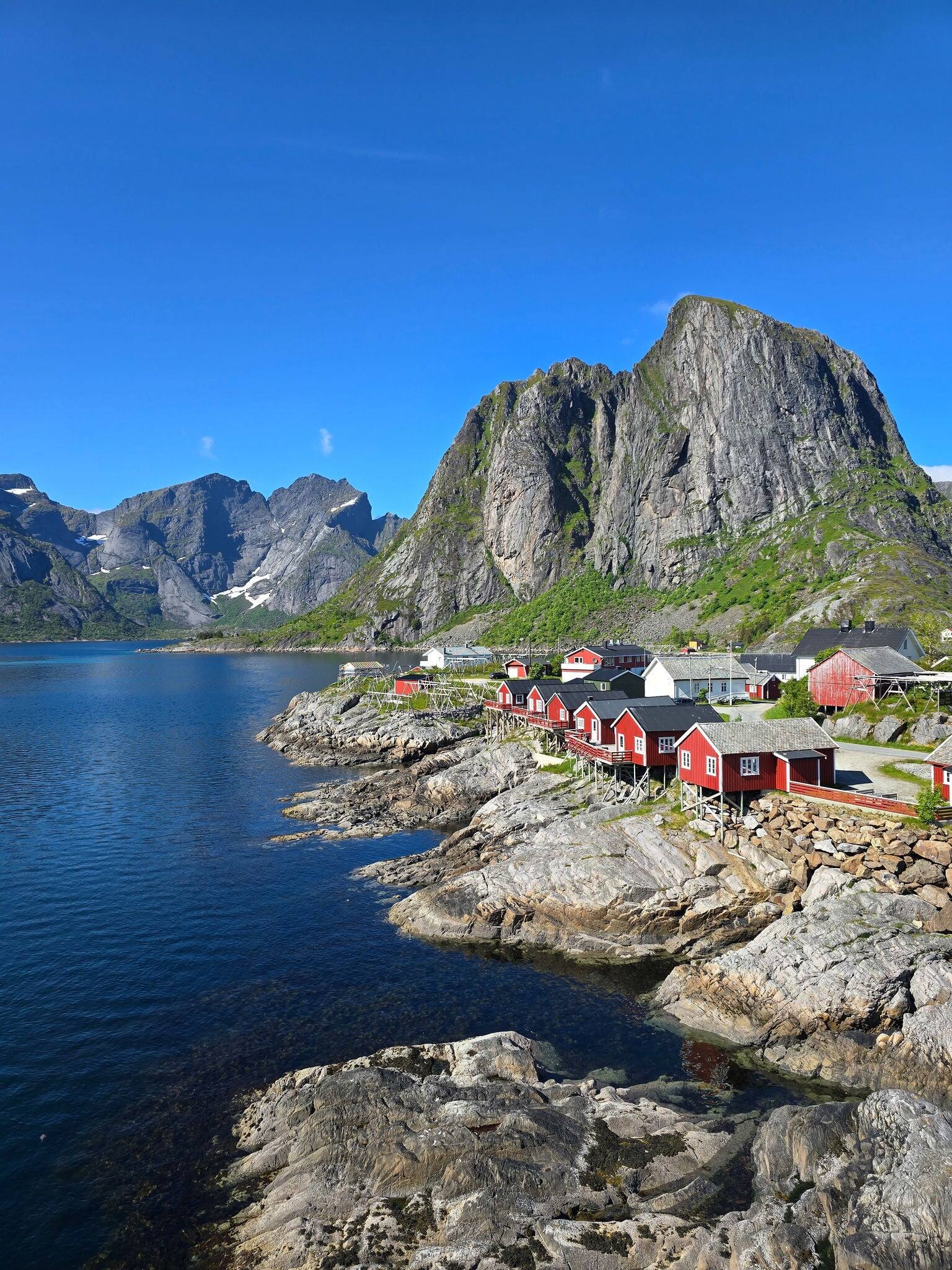 Nestled along the rocky coast, vibrant red cabins reflect against calm waters, surrounded by majestic mountains under a clear sky. Lofoten Norway