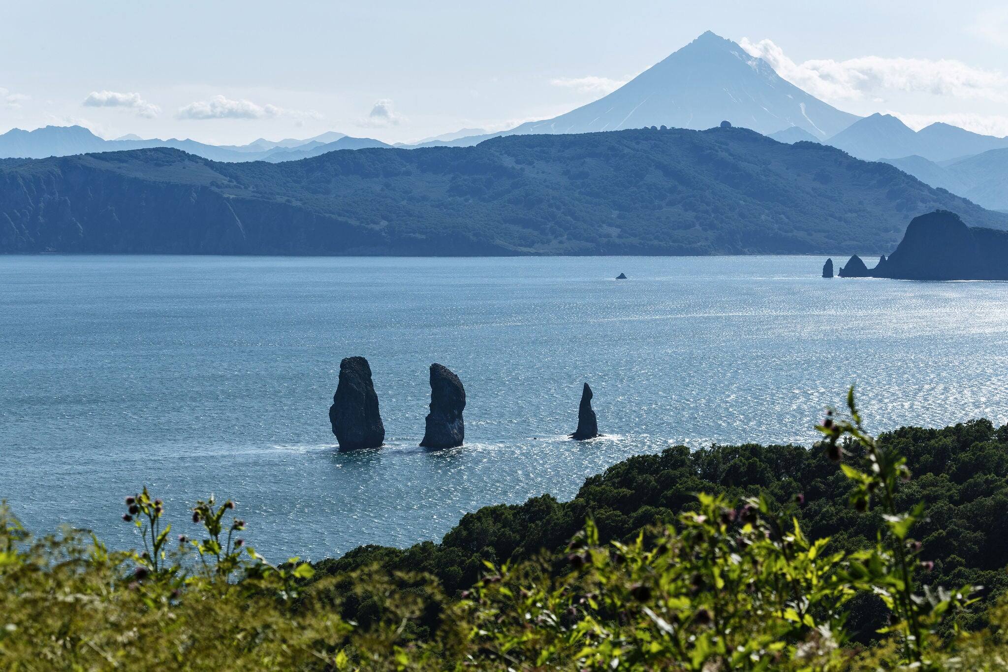 Beautiful summer landscape of Kamchatka Peninsula: Three Brothers Rocks in Avacha Bay (Pacific Ocean) and cone of Vilyuchinsky Volcano on other side of bay. Eurasia, Russian Far East, Kamchatka Region