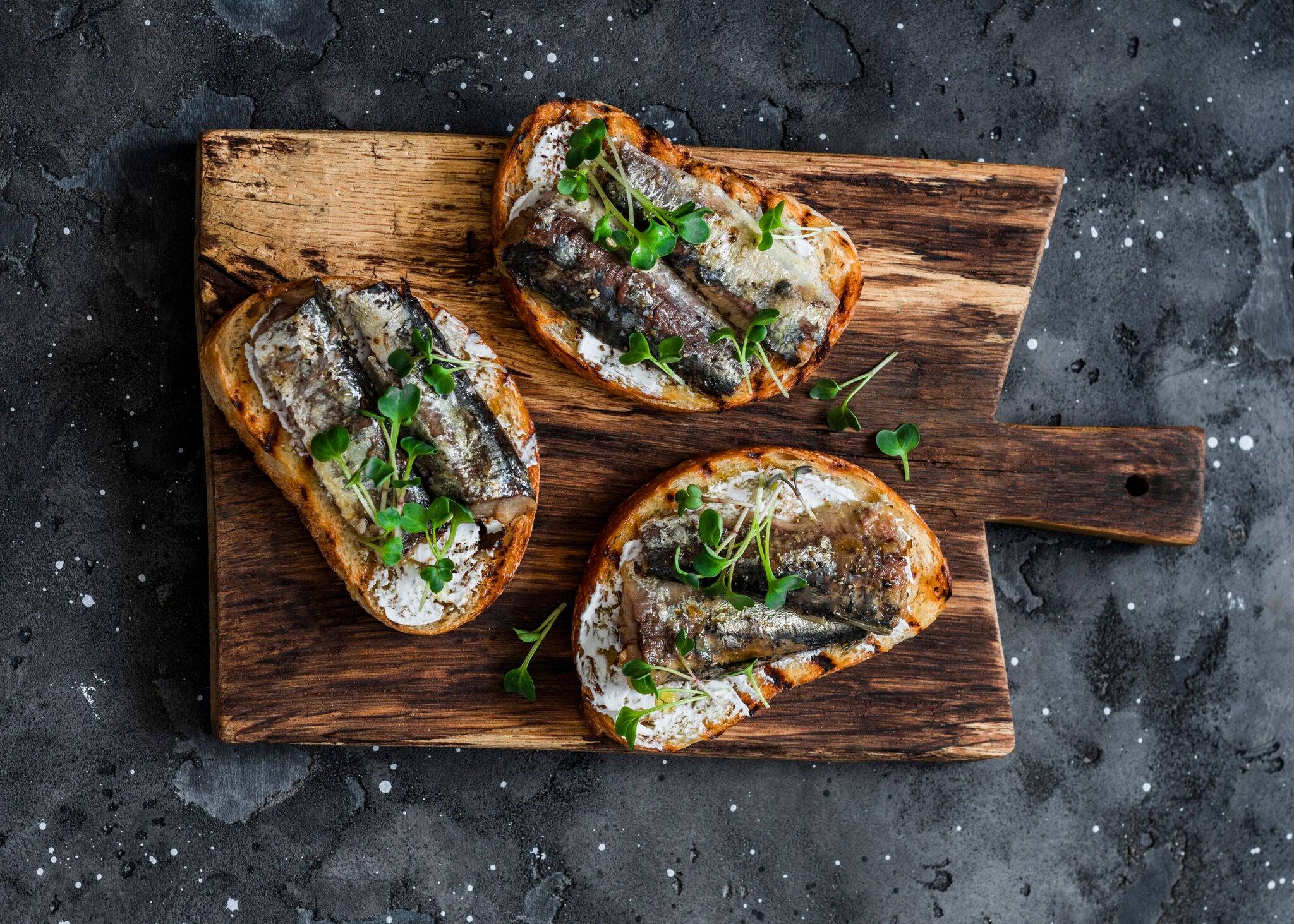 Grilled bread topped with sardines and micro greens on a rustic cutting board.