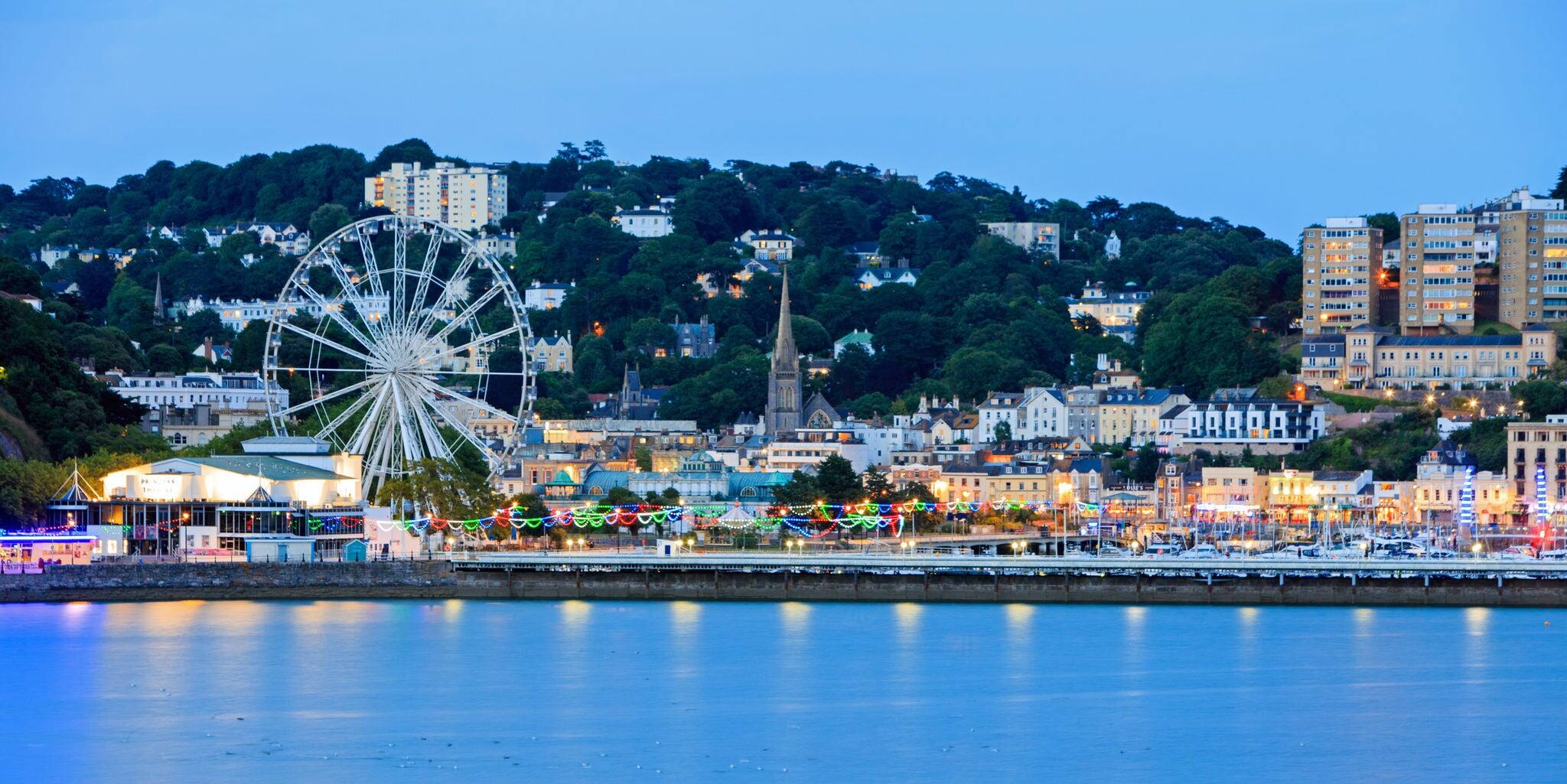 Torquay Promenade at dusk taken from a distance, showing the promenade illuminated with colourful lights that are reflecting in the calm sea. Devon, England