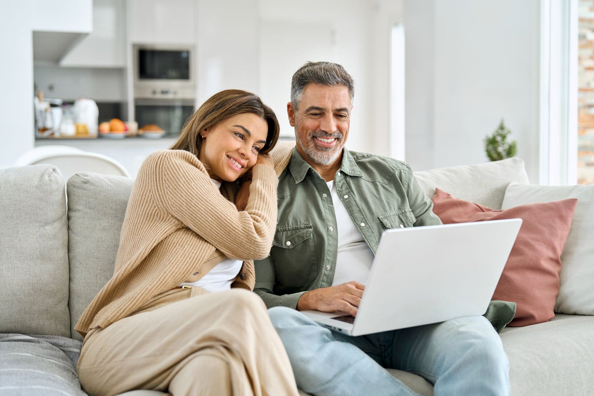 Happy middle aged couple using laptop relaxing on couch at home. Smiling mature man and woman looking at computer watching video, browsing or shopping in ecommerce store sitting on sofa in living room